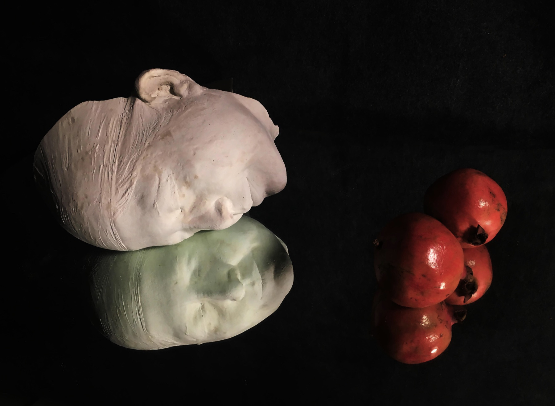 A plaster cast of a head beside small red pomegranates, against a black background